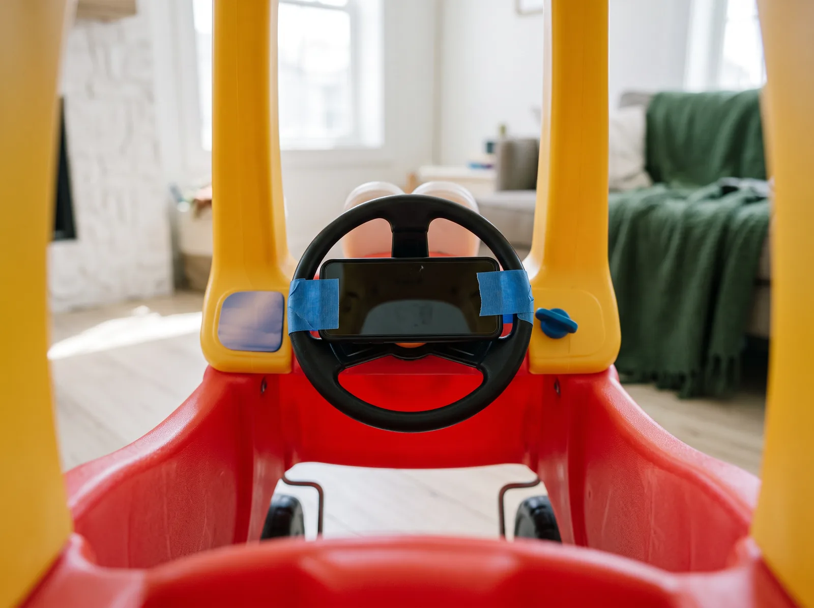 A phone taped to the steering wheel inside a red and yellow Little Tikes Cozy Coupe